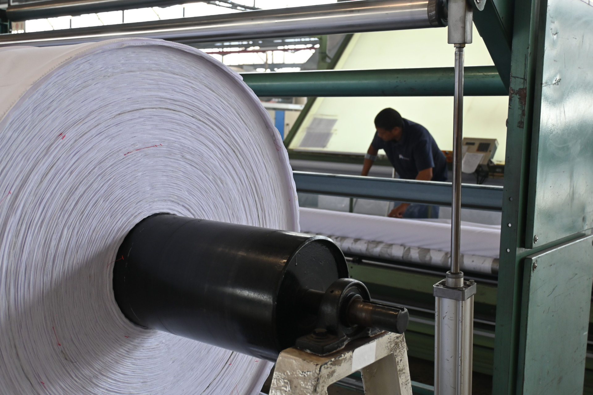 Large roll of finished white fabric on winder with operator inspecting in background